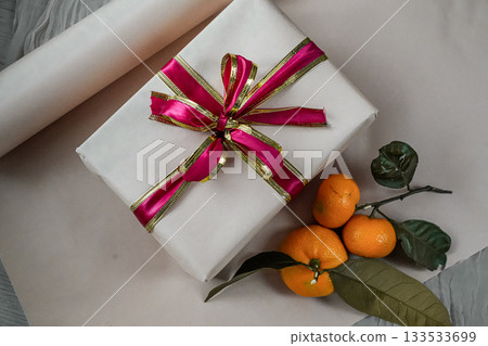 Medium view of a beige gift box decorated with a red ribbon, placed on a neutral background. Two fresh tangerines lie beside it, creating a warm and festive composition suitable for Christmas, New 133533699