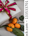 Close-up of a beige gift box decorated with a red ribbon, accompanied by two fresh tangerines on a neutral background. Minimal and festive composition ideal for Christmas, New Year, holiday, or 133533701