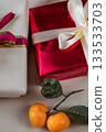 Close-up view of two gift boxes, one red and one beige, placed side by side on a neutral background. A fresh tangerine lies beside them, creating a festive and minimal holiday composition suitable for 133533703