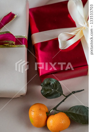 Close-up view of two gift boxes, one red and one beige, placed side by side on a neutral background. A fresh tangerine lies beside them, creating a festive and minimal holiday composition suitable for 133533703
