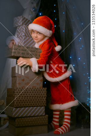 Smiling boy in Santa outfit standing by a Christmas tree with blue lights, embracing a large stack of wrapped gifts Smiling boy in Santa outfit standing by a Christmas tree with blue lights, embracing a large stack of wrapped gifts 133534026