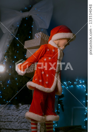 Child in Santa costume and hat near a festive tree with blue lights, carrying beautifully wrapped presents behind him 133534048