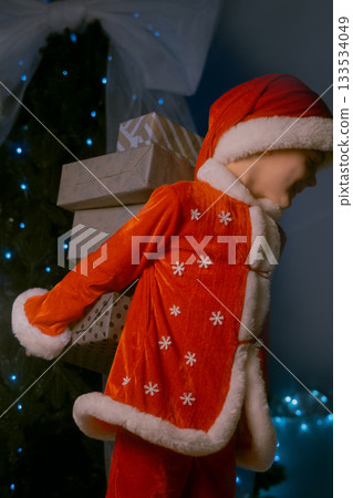 Boy dressed as Santa Claus standing by a Christmas tree with blue lights, holding several wrapped gifts behind his back 133534049