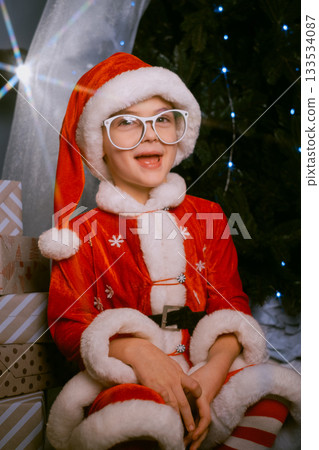 Child in a Santa costume on a sleigh by a festive tree with glowing blue lights, surrounded by wrapped presents 133534087