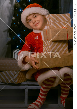 Boy dressed as Santa Claus sitting on sleigh near a Christmas tree with blue lights, holding many wrapped gifts indoors Boy dressed as Santa Claus sitting on sleigh near a Christmas tree with blue lights, holding many wrapped gifts indoors 133534152
