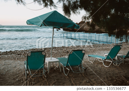 Turquoise loungers and umbrella on beach, pine tree shadow, pier leading into sea and strong waves Turquoise loungers and umbrella on beach, pine tree shadow, pier leading into sea and strong waves 133536677