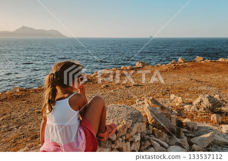 Little 6-year-old girl in striped dress and sunglasses sitting on rocks by the sea 133537112