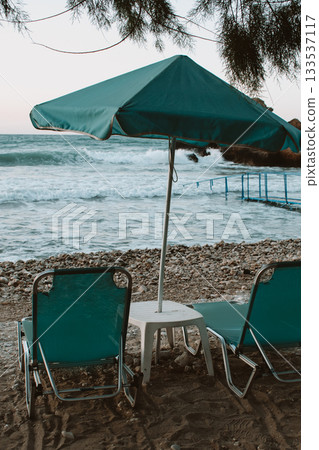 Beach scene at sunset with turquoise umbrella and sun loungers, small table between loungers, sea with small waves Beach scene at sunset with turquoise umbrella and sun loungers, small table between loungers, sea with small waves 133537117
