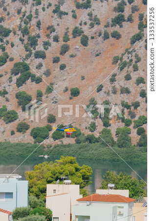 Blue and yellow firefighting helicopter flying over lake to collect water for extinguishing fire 133537256