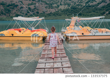 Georgioupoli, Crete, Greece. June 4, 2025. Little girl in pink dress walking on wooden pier to reach catamaran at Lake Kournas Georgioupoli, Crete, Greece. June 4, 2025. Little girl in pink dress walking on wooden pier to reach catamaran at Lake Kournas 133537375