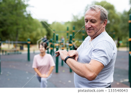 Elderly man looking at his smart bracelet in open-air sports area 133540043