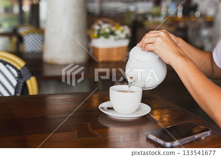 Female hand pouring from white ceramic teapot into a cup. Female hand pouring from white ceramic teapot into a cup. 133541677