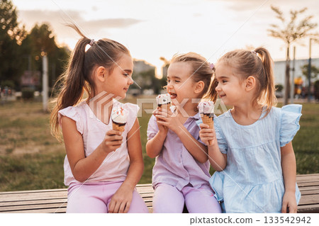 Three little girls eating ice cream cones in a park at sunset Three little girls eating ice cream cones in a park at sunset 133542942