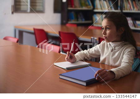 Young Girl Studying in Library, Writing in Notebook With Focused Expression 133544147