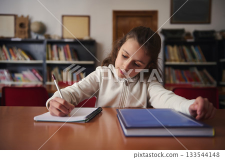 Young Girl Studies in Library, Writing in Notebook at Desk with Books Behind Young Girl Studies in Library, Writing in Notebook at Desk with Books Behind 133544148