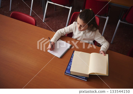 Young Student Writes Notes At Table With Open Book In Classroom Library 133544150