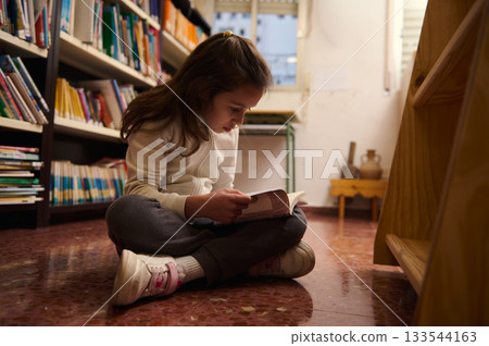 Curious Young Girl Reading a Book in a Quiet Library Aisle 133544163