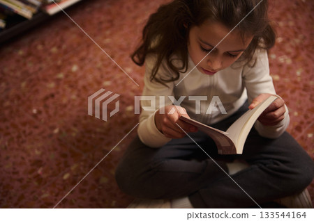 Young Girl Reading a Book While Sitting Cross-Legged on a Carpet in a Library 133544164