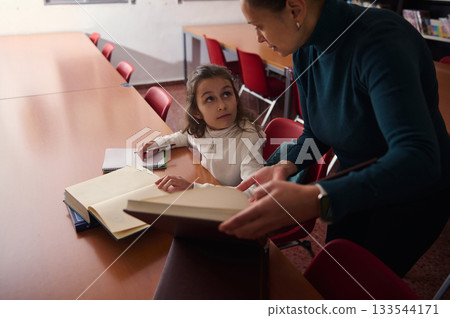 Young Girl Receives Help From Teacher Reading Books In Library Classroom Setting 133544171