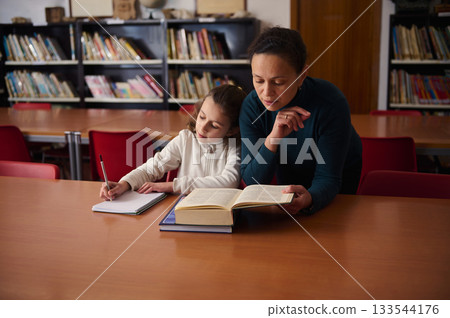 Mother and Child Studying Together in the Library, Reading a Book and Writing Notes 133544176