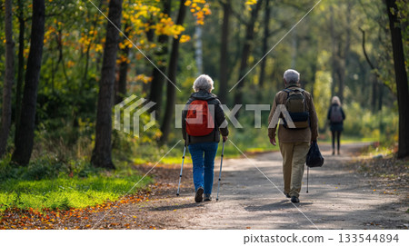 Two seniors with backpacks enjoy a leisurely walk on a path in a park, surrounded by the greenery of early fall. 133544894