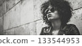 A fashion-forward woman in round sunglasses and a leather jacket leans against a wall, exuding confidence and coolness in a monochrome image. 133544953
