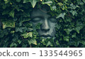 A stone face sculpture emerges from a bed of ivy, its features softly obscured by the green leaves, blending art with nature. 133544965