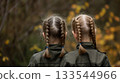 The back view of twin girls wearing green jackets, their hair styled in identical braids, against a backdrop of autumn leaves. 133544966