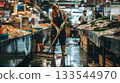 A market worker diligently mops the floor at a bustling fish market, ensuring cleanliness amidst the daily commerce. 133544970
