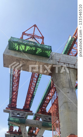 Concrete bridge pier and red steel gantry construction with green safety netting against blue sky, dynamic industrial scene 133546510