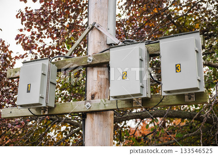 Numbered electrical junction boxes mounted on a wooden utility pole with cables and autumn foliage in the background, part of outdoor power infrastructure. Numbered electrical junction boxes mounted on a wooden utility pole with cables and autumn foliage in the background, part of outdoor power infrastructure. 133546525