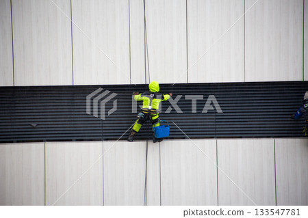 Man in bright reflective suit works on ropes with safety harness. Industrial climber performs difficult, risky work. Cleaning building ventilation system with help of professional climbers 133547781