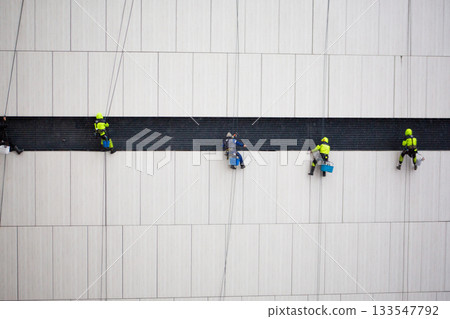 Industrial climbers on belay rope on the side of building. Performing dangerous work. Men with specialized equipment hang from the wall of building while climbers work Industrial climbers on belay rope on the side of building. Performing dangerous work. Men with specialized equipment hang from the wall of building while climbers work 133547792