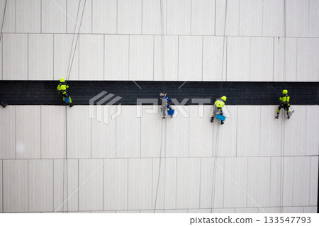 Construction crew of climbers performs dangerous task. Men in protective suits sit on seat suspended by ropes at great height. Industrial climbers work on the wall of building 133547793