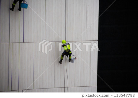 Climber in reflective protective suit uses ropes and safety equipment to descend the building facade during work. Concept of working at height and in challenging weather conditions Climber in reflective protective suit uses ropes and safety equipment to descend the building facade during work. Concept of working at height and in challenging weather conditions 133547798