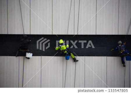 Industrial climbers wipe down building facade element with cloth. Climbers work at height. Safety equipment is essential for dangerous, life threatening work Industrial climbers wipe down building facade element with cloth. Climbers work at height. Safety equipment is essential for dangerous, life threatening work 133547805