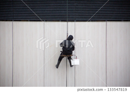 Industrial climber in warm clothing sits on bench with ropes in bad weather. Dangerous work at height. Industrial mountaineering concept. The climber hangs from the wall of a building Industrial climber in warm clothing sits on bench with ropes in bad weather. Dangerous work at height. Industrial mountaineering concept. The climber hangs from the wall of a building 133547819