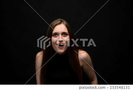 Surprised young woman with wide eyes reacting emotionally in dramatic studio portrait on dark background Surprised young woman with wide eyes reacting emotionally in dramatic studio portrait on dark background 133548131