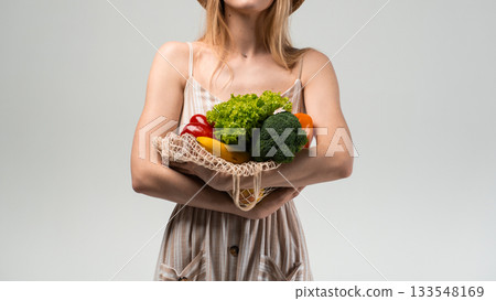Woman in striped dress holding eco mesh bag with fresh vegetables in hands on light background Woman in striped dress holding eco mesh bag with fresh vegetables in hands on light background 133548169