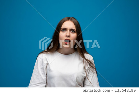 Surprised young woman reacting with wide eyes and open mouth in a bright blue studio environment 133548199