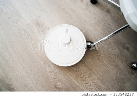 Robotic vacuum navigating near office chair wheel on wooden flooring during automated cleaning routine at home Robotic vacuum navigating near office chair wheel on wooden flooring during automated cleaning routine at home 133548237