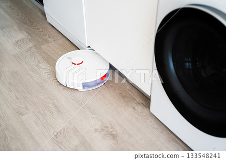 Robot vacuum cleaner moving near washing machine on wooden floor inside bright modern interior Robot vacuum cleaner moving near washing machine on wooden floor inside bright modern interior 133548241