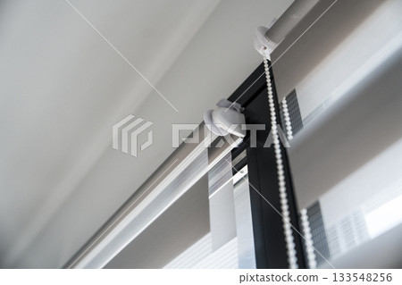 Detail of mounted zebra roller blinds with chain mechanism and striped fabric layers on interior window Detail of mounted zebra roller blinds with chain mechanism and striped fabric layers on interior window 133548256