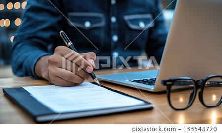 Individual is focused on writing notes with a pen while seated at a desk with a laptop and glasses nearby, creating a warm atmosphere Individual is focused on writing notes with a pen while seated at a desk with a laptop and glasses nearby, creating a warm atmosphere 133548720