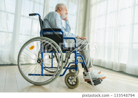 Sad senior man sitting in wheelchair looking out window in retirement home hospital. Old paralyzed man in chair for people with disability feel depressed. Lonely elderly sick man sitting on wheelchair 133548794
