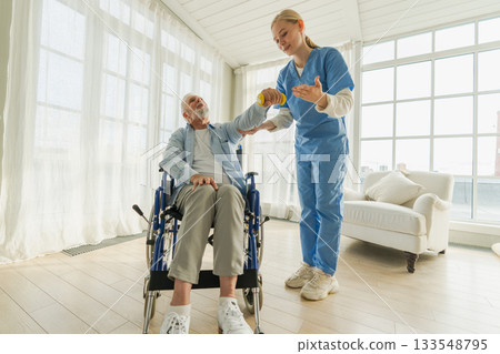 Support rehabilitation healthcare. Nurse helps old man patient in wheelchair picking up dumbbells. Paralyzed man in chair for people with disability doing sports exercise with nurse. Medical caregiver 133548795