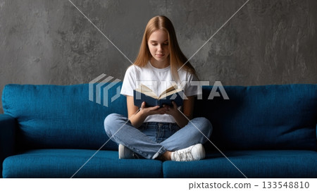 Young woman sits on blue couch reading a book in a cozy living room on a quiet afternoon 133548810