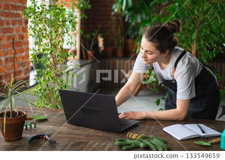 Female florist gardener working in botanical store greenhouse using laptop order plants communicating on Internet with customer. Small business owner working at flower shop. Running of own business Female florist gardener working in botanical store greenhouse using laptop order plants communicating on Internet with customer. Small business owner working at flower shop. Running of own business 133549659