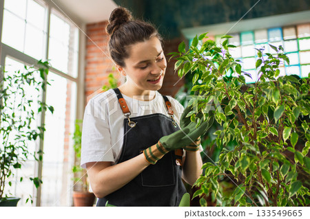 Running of own business. Woman florist wearing apron taking care of green plants in botanical store. Happy small business owner working at flower shop standing surrounded by plants. Small business 133549665