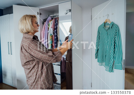 Middle aged Woman taking photo of shirt using smartphone for selling or donating her clothes. Decluttering , Sorting Clothes, And Cleaning Up wardrobe. Reuse, second-hand concept. Conscious consumer. Middle aged Woman taking photo of shirt using smartphone for selling or donating her clothes. Decluttering , Sorting Clothes, And Cleaning Up wardrobe. Reuse, second-hand concept. Conscious consumer. 133550378